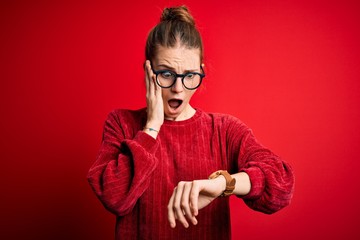 Young beautiful redhead woman wearing casual sweater over isolated red background Looking at the watch time worried, afraid of getting late