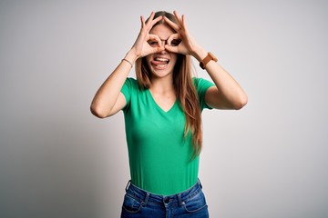 Young beautiful redhead woman wearing casual green t-shirt and glasses over white background doing ok gesture like binoculars sticking tongue out, eyes looking through fingers. Crazy expression.