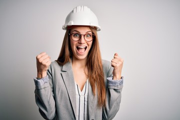 Young beautiful redhead architect woman wearing security helmet over white background celebrating surprised and amazed for success with arms raised and open eyes. Winner concept.
