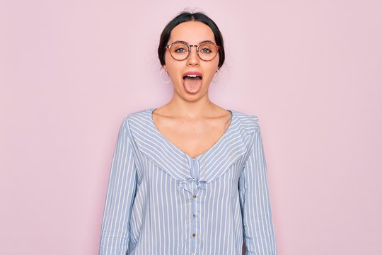 Young beautiful woman wearing casual striped shirt and glasses over pink background sticking tongue out happy with funny expression. Emotion concept.