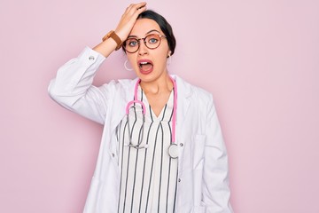 Beautiful doctor woman with blue eyes wearing coat and stethoscope over pink background surprised...