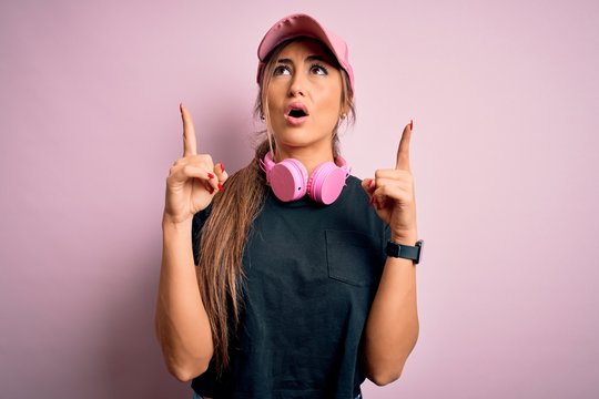 Young beautiful fitness sports woman wearing training cap and headphones over pink background amazed and surprised looking up and pointing with fingers and raised arms.