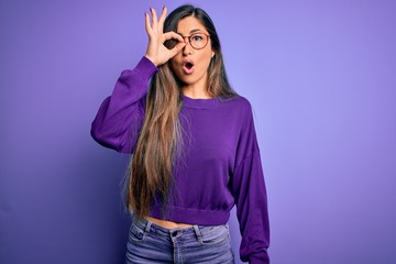 Naklejka premium Young beautiful smart woman wearing glasses over purple isolated background doing ok gesture shocked with surprised face, eye looking through fingers. Unbelieving expression.