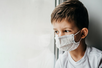 a little boy in a medical mask is sitting at home in quarantine because of coronavirus and covid -19 and looks out the window.