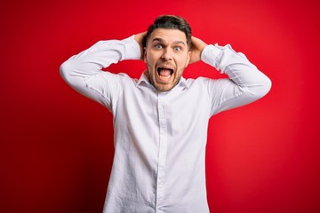 Young business man with blue eyes wearing elegant shirt standing over red isolated background Crazy and scared with hands on head, afraid and surprised of shock with open mouth