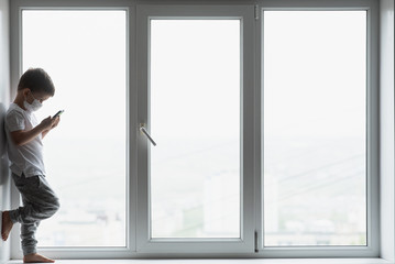 Fototapeta premium A small child in a medical mask sits quarantined at home on a window with a phone in his hands.Prevention of coronavirus and Covid - 19