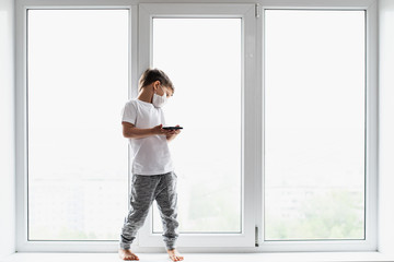 A little boy in a medical mask is sitting on the window of the house in quarantine with a phone in his hands.Prevention of coronavirus and Covid - 19