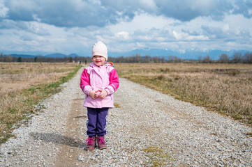 Naklejka na ściany i meble Cute girl wearing pink jacket standing on road running through fields.