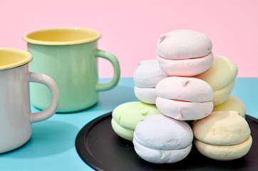 Top view of pastel colored marshmallow on black plate and two empty cups on a blue pink background. Minimalism photo for your design