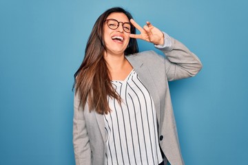 Young hispanic business woman wearing glasses standing over blue isolated background Doing peace symbol with fingers over face, smiling cheerful showing victory