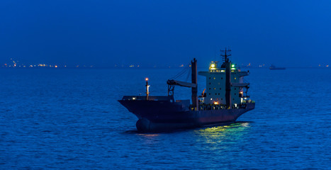 Container cargo ship with navigation lights anchored in outer anchorage of Singapore at golden hour.