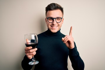 Young handsome caucasian man drinking an alcoholic glass of red wine over isolated background surprised with an idea or question pointing finger with happy face, number one