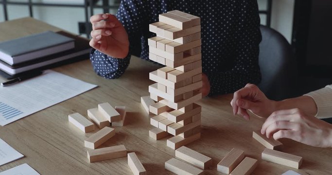 Two business women play jenga game in office. Female business partners hands take blocks out of wooden tower. Risky solutions in company building strategy plan, help in teamwork concept. Close up view