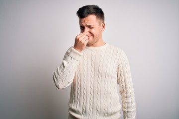 Young handsome caucasian man wearing casual winter sweater over white isolated background smelling something stinky and disgusting, intolerable smell, holding breath with fingers on nose. Bad smell