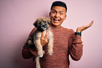 Young handsome latin man holding cute puppy pet over isolated pink background very happy and excited, winner expression celebrating victory screaming with big smile and raised hands