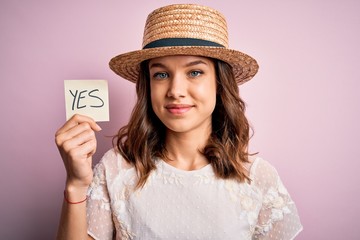 Young blonde girl wearing a hat holding paper note with yes word as positive message with a happy face standing and smiling with a confident smile showing teeth