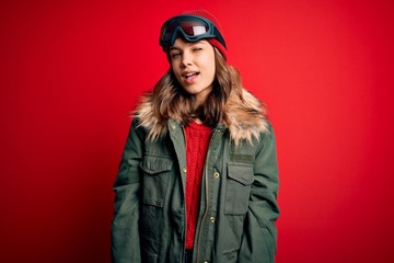 Young blonde girl wearing ski glasses and winter coat for ski weather over red background winking...