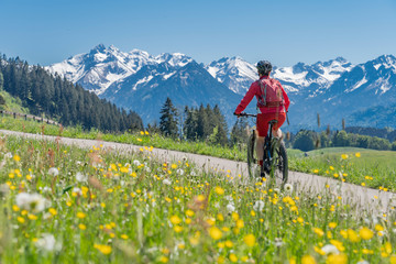 pretty senior woman riding her electric mountain on warm spring day with snoe capped bike in the Iller valley near Oberstdorf with snoe capped Allgau High Alps in background, Allgau Alps, Bavaria, 