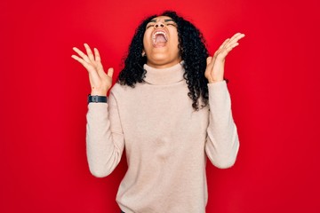 Young african american curly woman wearing casual turtleneck sweater over red background celebrating mad and crazy for success with arms raised and closed eyes screaming excited. Winner concept