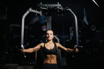 Fototapeta na wymiar Middle shot portrait of sportive young woman with perfect athletic body wearing black sportswear using training simulator for pumping back muscles during sport workout training in modern dark gym.