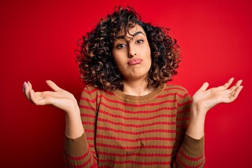 Young beautiful curly arab woman wearing casual striped sweater standing over red background clueless and confused expression with arms and hands raised. Doubt concept.