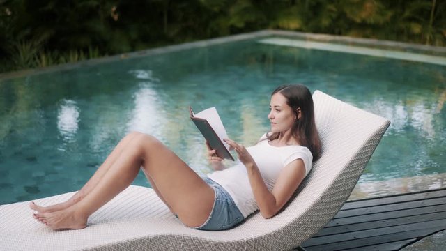 Young woman in summer clothes lies on deck chair and reads book near pool