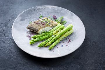 Gebratenes Gourmet Skrei Kabeljau Fisch Filet mit grünem Spargel und Salat angeboten als closeup auf einem Modern Design Teller mit Textfreiraum 