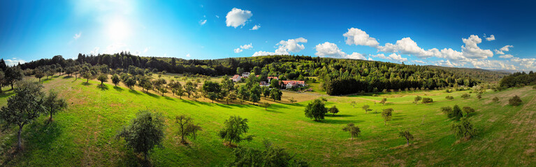 Fototapeta na wymiar Wide aerial panorama view of a nice rural landscape with green meadows on hills, trees and a few houses, deep blue sky, the sun and white clouds