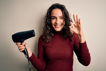 Young beautiful woman with curly hair using hair dryer over isolated white background doing ok sign with fingers, excellent symbol