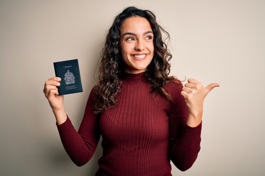 Young beautiful tourist woman with curly hair on vacation holding canadian canada passport pointing and showing with thumb up to the side with happy face smiling