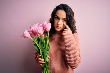 Young beautiful romantic woman with curly hair holding bouquet of pink tulips Pointing to the eye watching you gesture, suspicious expression