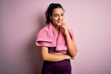 Young beautiful sportswoman with curly hair doing sport using towel over pink background looking confident at the camera with smile with crossed arms and hand raised on chin. Thinking positive.