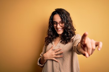 Beautiful woman with curly hair wearing striped shirt and glasses over yellow background laughing at you, pointing finger to the camera with hand over body, shame expression