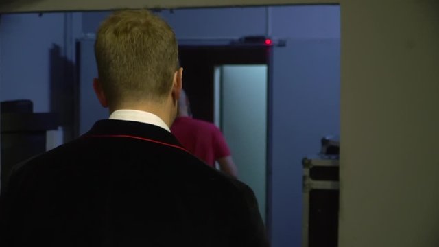 Two men follow each other down the hall. A guy in a black suit with a red stripe. A bald man in a red Polo.
