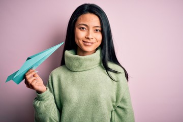 Young beautiful chinese woman holding paper plane standing over isolated pink background with a happy face standing and smiling with a confident smile showing teeth