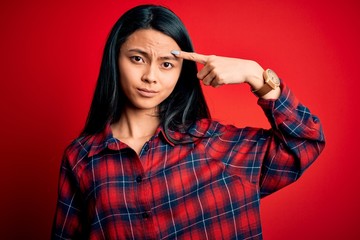 Young beautiful chinese woman wearing casual shirt over isolated red background pointing unhappy to pimple on forehead, ugly infection of blackhead. Acne and skin problem