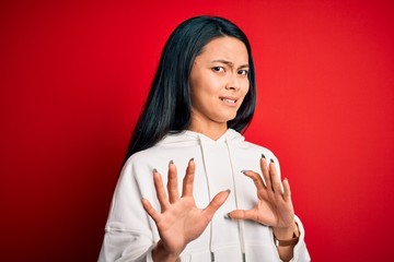 Young beautiful chinese sporty woman wearing sweatshirt over isolated red background disgusted expression, displeased and fearful doing disgust face because aversion reaction. With hands raised