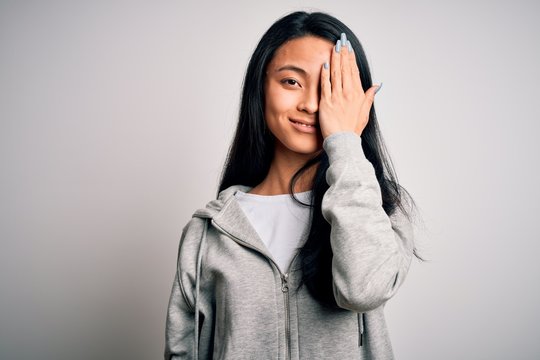 Young beautiful chinese sporty woman wearing sweatshirt over isolated white background covering one eye with hand, confident smile on face and surprise emotion.
