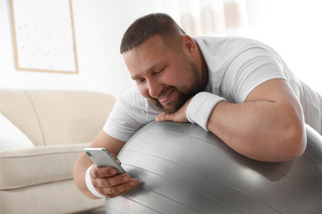 Lazy overweight man using smartphone while lying on exercise ball at home