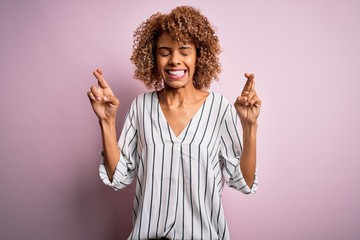 Beautiful african american woman with curly hair wearing striped t-shirt over pink background gesturing finger crossed smiling with hope and eyes closed. Luck and superstitious concept.