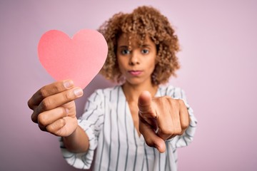 Young african american romantic woman with curly hair holding paper heart shape pointing with finger to the camera and to you, hand sign, positive and confident gesture from the front