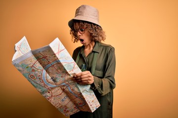 African american tourist woman wearing explorer hat and water canteen holding city map scared in...