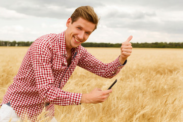 Young handsome man standing in wheat field holding mobile and showing hand ok sign