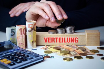 Verteilung. Männliche Hand stapelt Geld-Turm (Euro). Begriff an Baustein. Münzen, Scheine &...