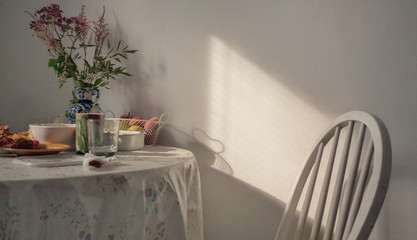 Table with a tablecloth, flowers and fruits. White chair. Sunlight. Homely atmosphere.