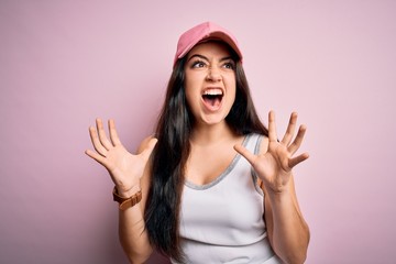Obraz na płótnie Canvas Young brunette woman wearing casual sport cap over pink background crazy and mad shouting and yelling with aggressive expression and arms raised. Frustration concept.