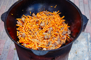 Preparation of national uzbek dish pilaf, pilaw, plov, carrot with meat in big pan. Cooking process, open fire. Cooking in a cauldron on fire. Stir slowly with a skimmer. Addition of small cut carrots