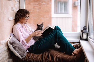 girl in glasses reads a book near the window. Young woman is relaxing on the balcony with her kitten