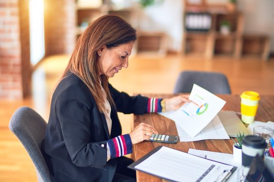 Middle age beautiful businesswoman smiling happy and confident. Sitting on chair working in a desk holding documents with diagrams at the office