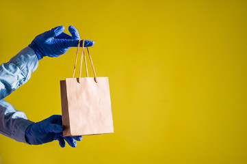 Female hands in gloves holds a small brown paper bag with handles on a yellow background. Safe food...